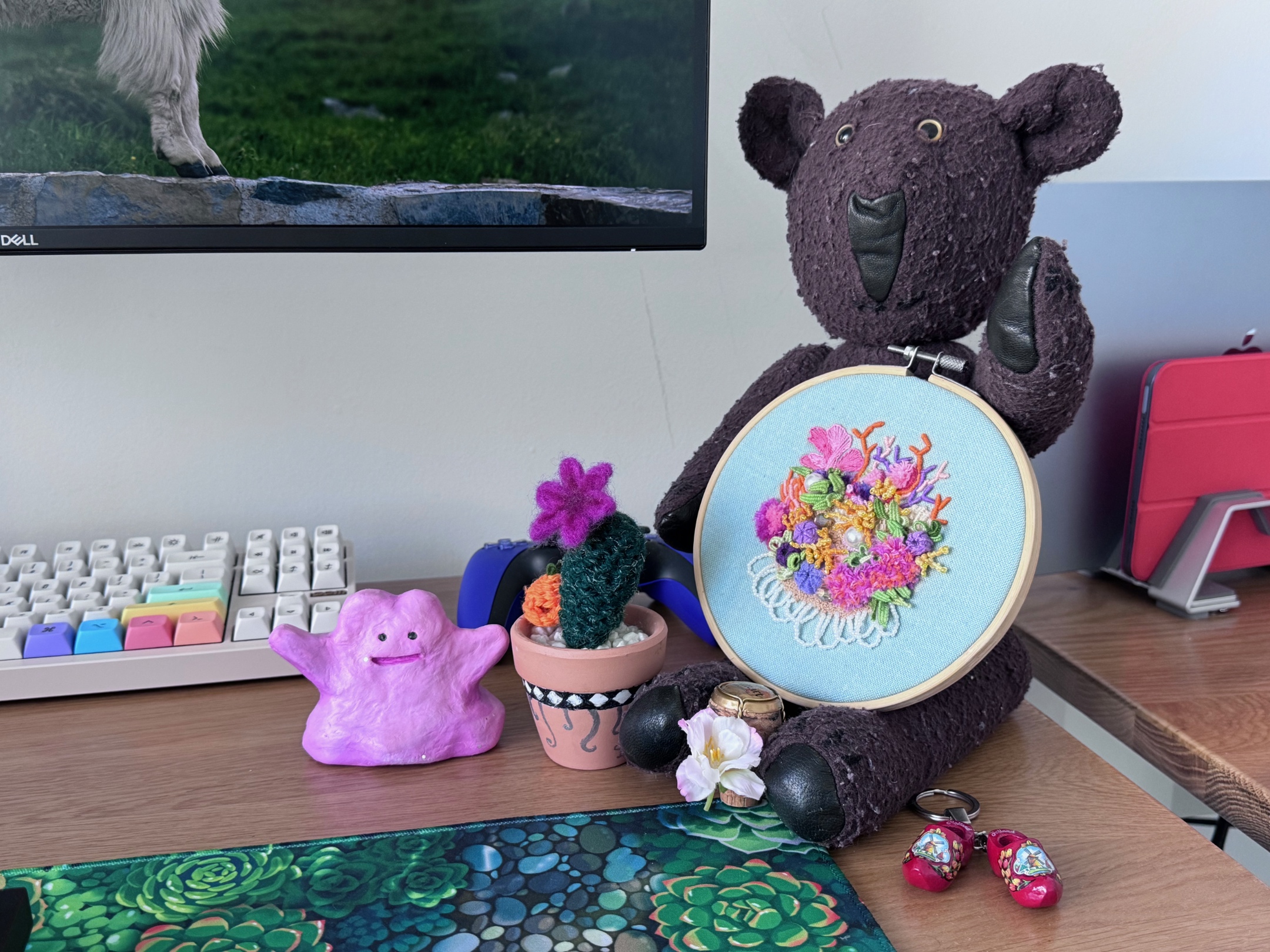 A teddy bear holding an embroidery, and sitting next to a keychain pair of crocs, a crocheted cactus, and a sculpted pink model of the Pokémon Ditto