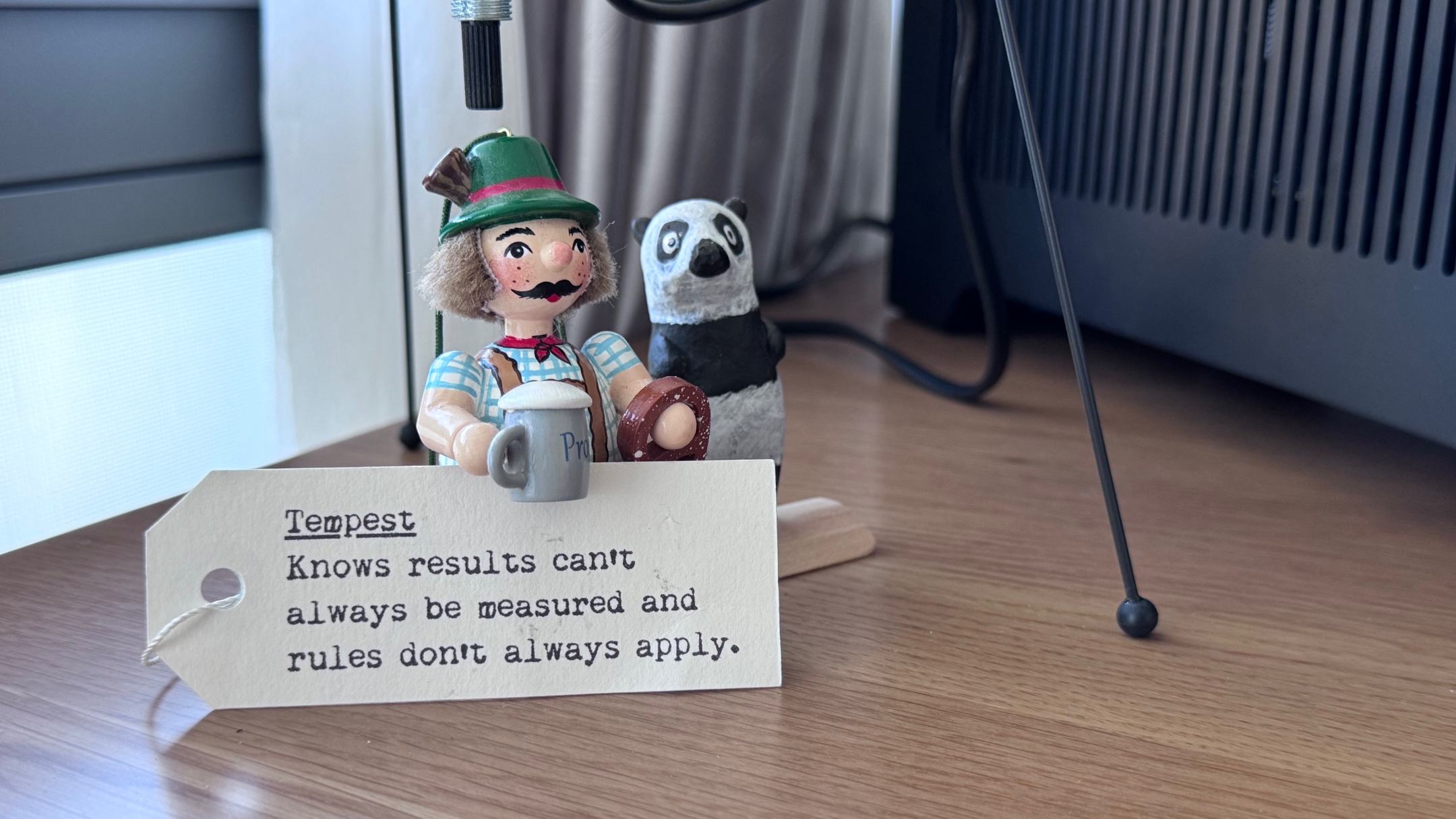 A wooden German figurine, and a stylised panda bear, standing under a lamp. The German is wearing lederhosen, holding a pretzel in one hand, and a beer stein in the other. Leaning against him is a tag that read: Tempest: Knows results can't always be measured and rules don't always apply.